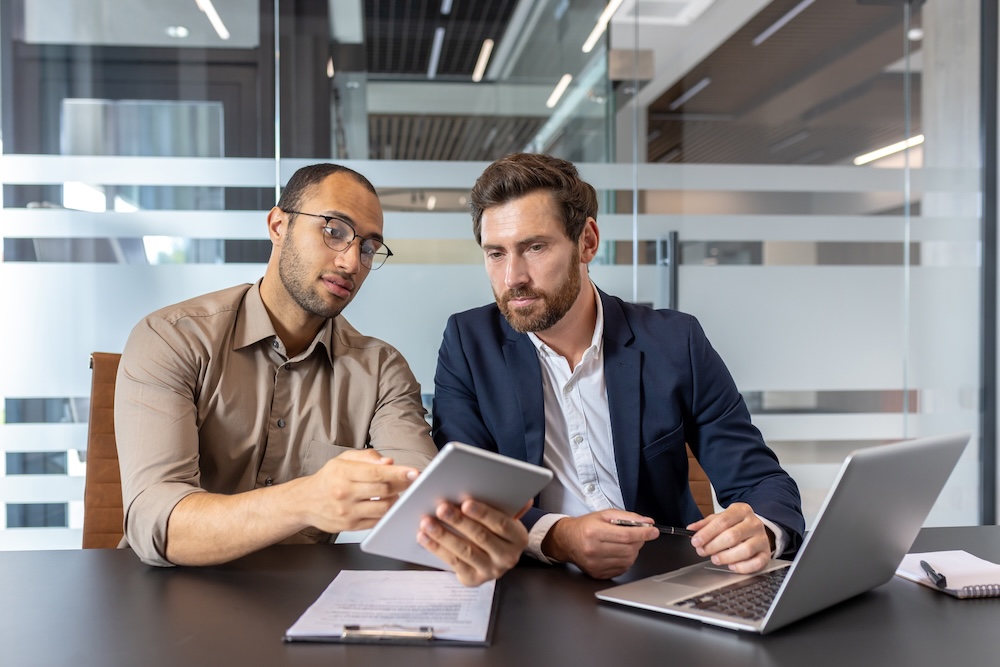 Two businessmen reviewing a tablet and laptop, discussing business strategies in a modern office setting.