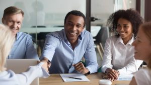 Smiling manager handshake greeting client at group negotiation, company representative shake hand of new partner get respect make business deal at diverse team meeting 