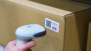 Handheld barcode scanner reading a shipping label with a QR code and traditional 1D barcode on a cardboard package.